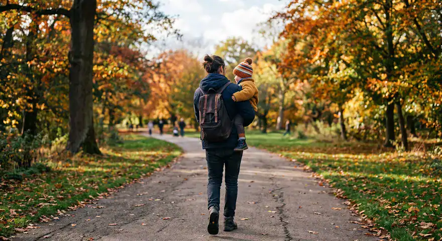 full-length shot from behind of a parent walking down a paved path in a park, carrying a young child on their hip. Neither face is visible. The parent wears a dark jacket, jeans, and a backpack, while the child wears a yellow coat and a striped knit beanie. Vibrant autumn trees line both sides of the path.
